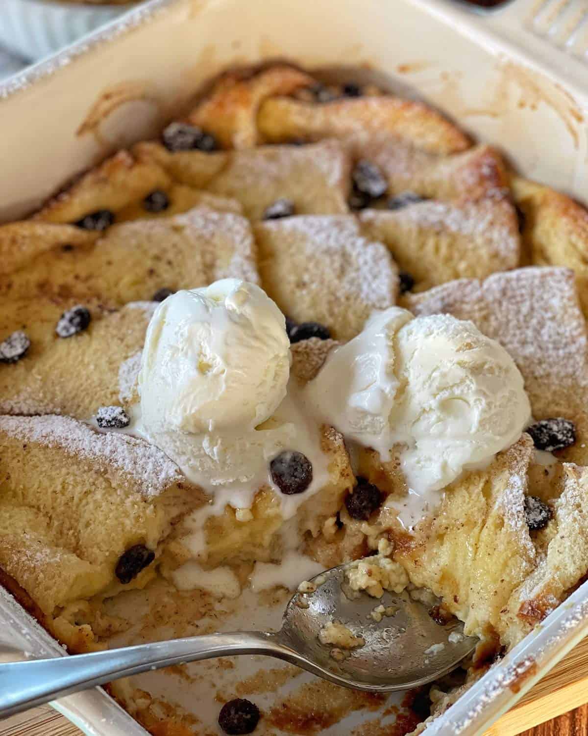 Bread and Butter Pudding A baking dish with bread pudding topped with raisins, powdered sugar, and two scoops of melting vanilla ice cream, with a serving spoon inside.