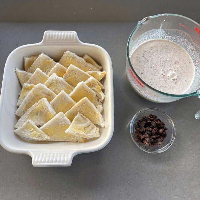 Bread and Butter Pudding A baking dish with buttered bread triangles, a measuring cup of milk mixture, and a small bowl of raisins on a gray countertop.