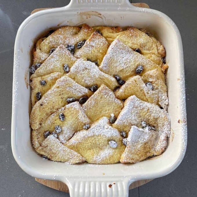 Bread and Butter Pudding A baked bread and butter pudding topped with raisins and dusted with powdered sugar in a square white ceramic dish.