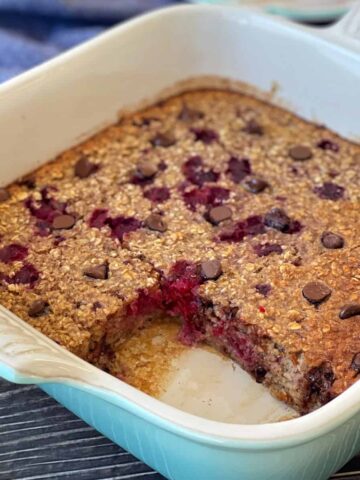A baked oatmeal dish with visible chocolate chips and berries, with one square piece removed, in a light blue baking pan on a dark surface.