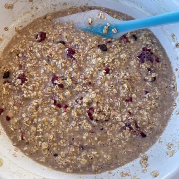 Banana and Raspberry Baked Oats A close-up of a mixing bowl containing uncooked oatmeal, milk, and visible berries, stirred with a blue spatula.