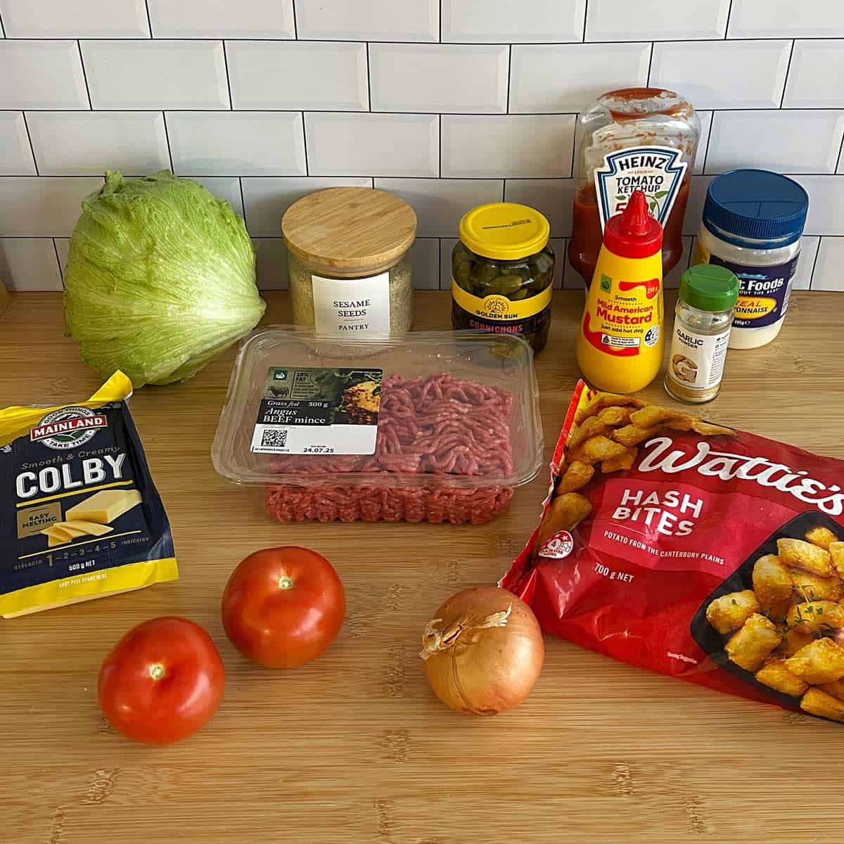 Hash Bite Burger Bowls A selection of ingredients on a wooden surface, including lettuce, cheese, tomatoes, ground beef, onion, condiments, seasonings, and a bag of frozen hash bites.