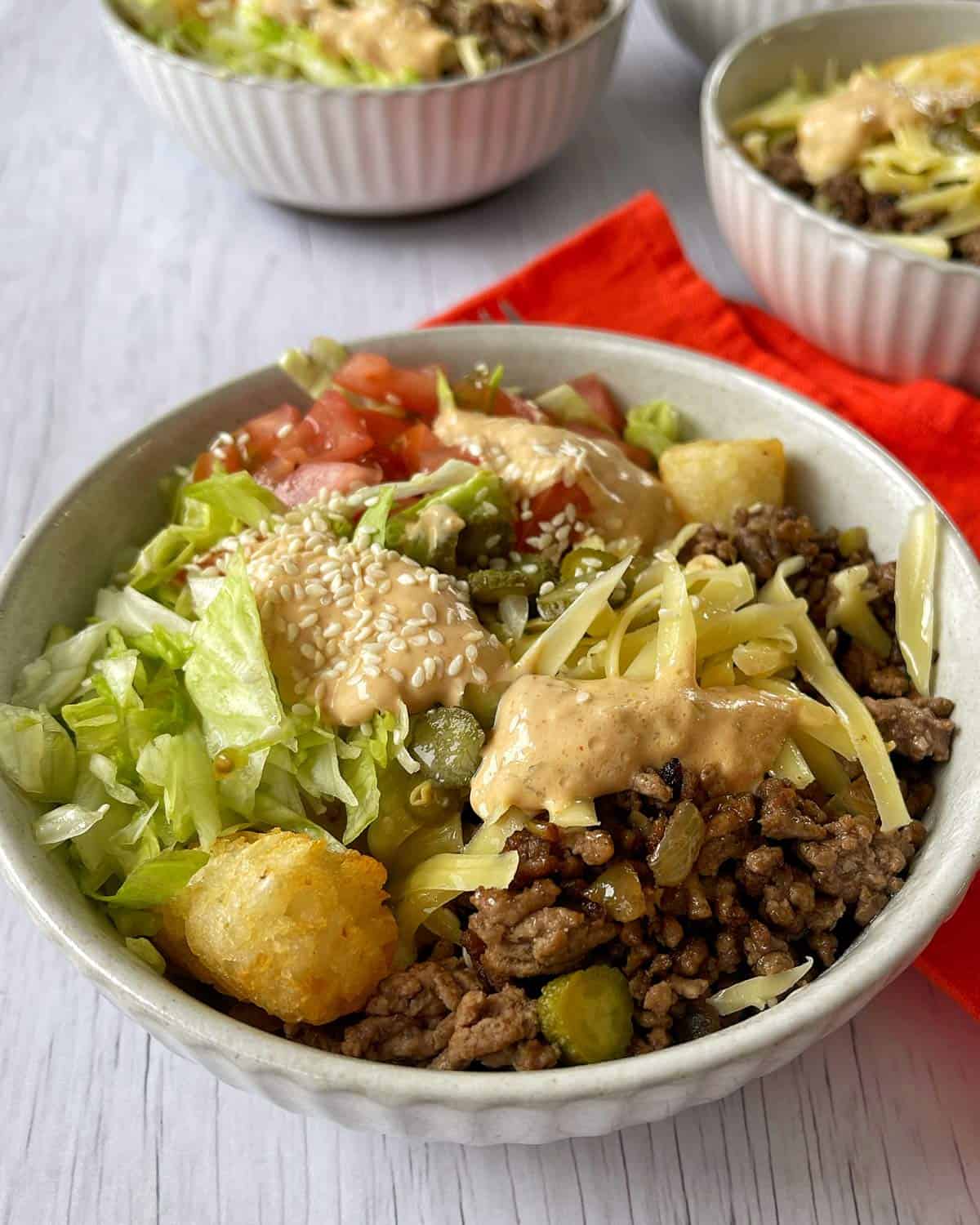 Hash Bite Burger Bowls A bowl filled with ground beef, shredded lettuce, cheese, diced tomatoes, tater tots, pickles, sesame seeds, and a creamy sauce, placed on a white surface with an orange napkin.