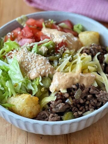 A bowl filled with ground beef, shredded lettuce, chopped tomatoes, shredded cheese, tater tots, pickles, sesame seeds, and a drizzle of creamy sauce.