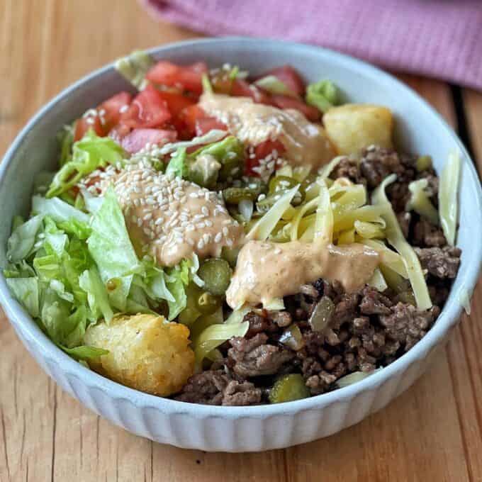 Hash Bite Burger Bowls A bowl filled with ground beef, shredded lettuce, chopped tomatoes, shredded cheese, tater tots, pickles, sesame seeds, and a drizzle of creamy sauce.