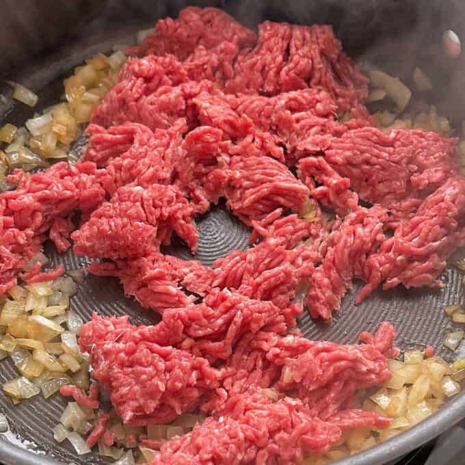 Hash Bite Burger Bowls Raw ground beef and diced onions are cooking in a frying pan. Steam is rising as the meat begins to brown and the onions appear translucent.