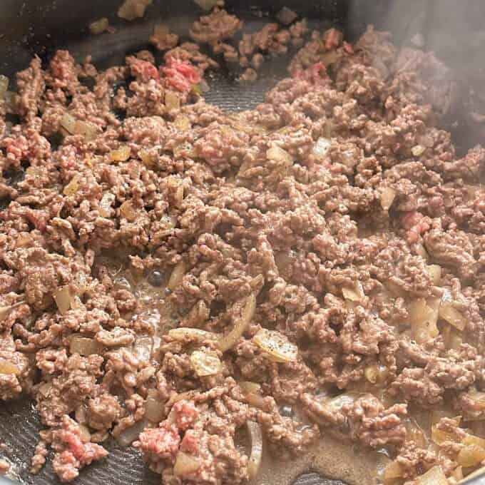 Hash Bite Burger Bowls Ground beef and chopped onions are being cooked in a skillet, with some pieces still slightly pink and others browned. Steam rises from the mixture.