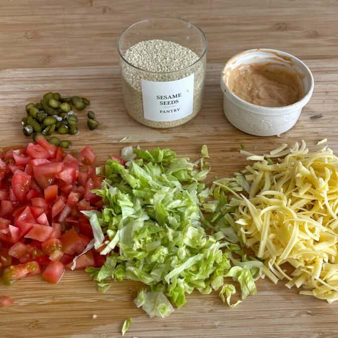 Hash Bite Burger Bowls Chopped capers, diced tomatoes, shredded lettuce, grated cheese, sesame seeds in a jar, and a small bowl of sauce arranged on a wooden surface.