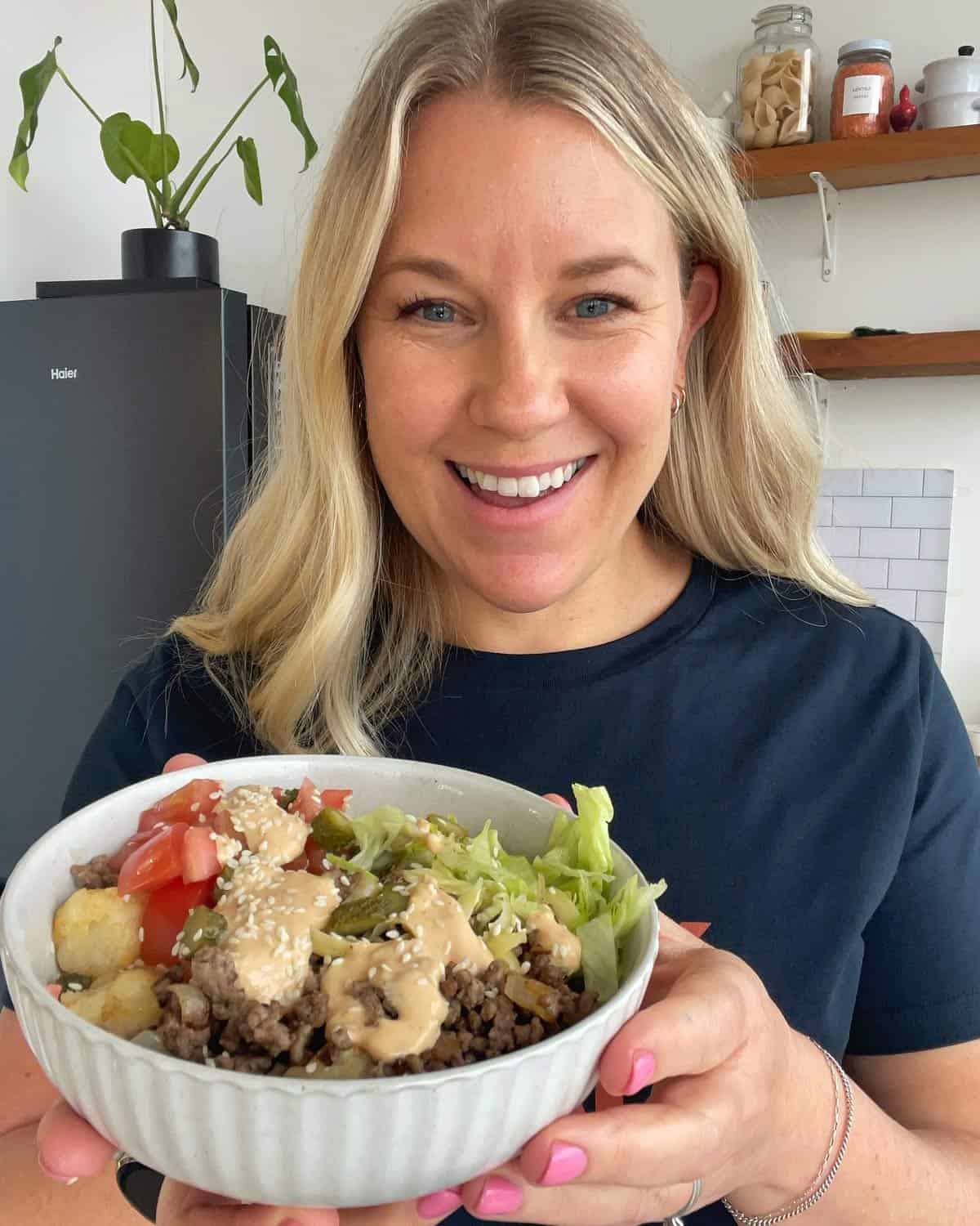 Hash Bite Burger Bowls A woman smiles while holding a bowl filled with ground meat, lettuce, tomato, sauce, and sesame seeds in a kitchen.
