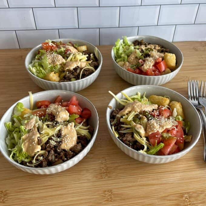 Hash Bite Burger Bowls Four bowls filled with ground meat, shredded lettuce, diced tomatoes, tater tots, shredded cheese, and sauce, arranged on a wooden surface in front of a tiled wall.