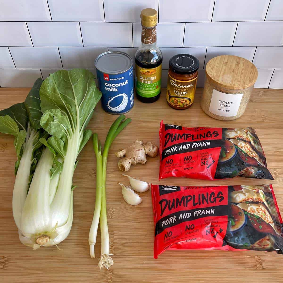 Thai Curry Dumpling Bake Fresh bok choy, green onions, garlic, ginger, canned coconut milk, gluten-free soy sauce, yellow curry paste, sesame seeds, and two packs of pork and prawn dumplings on a wooden surface.