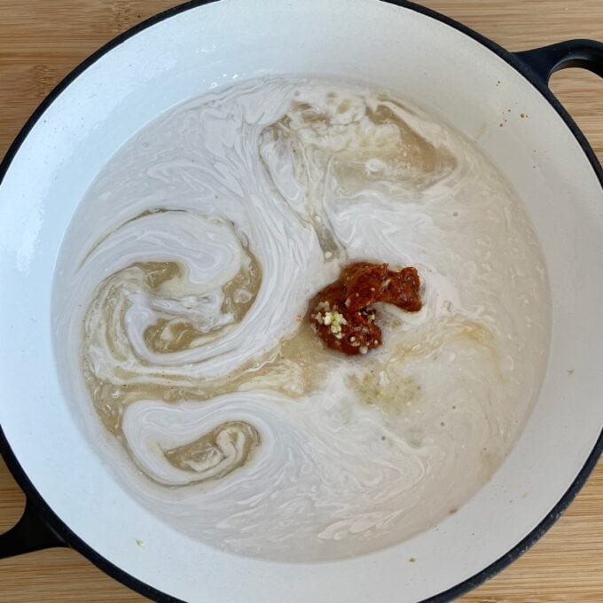 Thai Curry Dumpling Bake A white pot containing coconut milk, brown liquid, minced garlic, and red chili paste being mixed together.