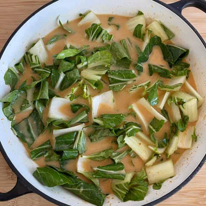 Thai Curry Dumpling Bake A white pan filled with chopped bok choy simmering in a light brown, creamy sauce, placed on a wooden surface.