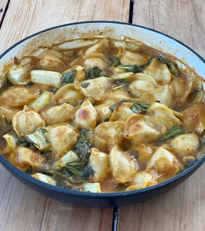 Thai Curry Dumpling Bake A round baking dish filled with cooked pasta shells, leafy greens, and a tomato-based sauce, placed on a wooden surface.