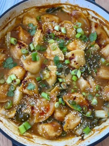 A pot filled with Thai Curry Dumpling Bake in a brown sauce, garnished with chopped green onions, sesame seeds, and cilantro.