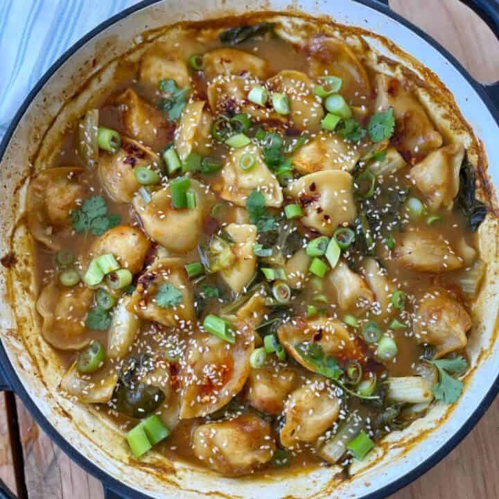 A pot filled with Thai Curry Dumpling Bake in a brown sauce, garnished with chopped green onions, sesame seeds, and cilantro.