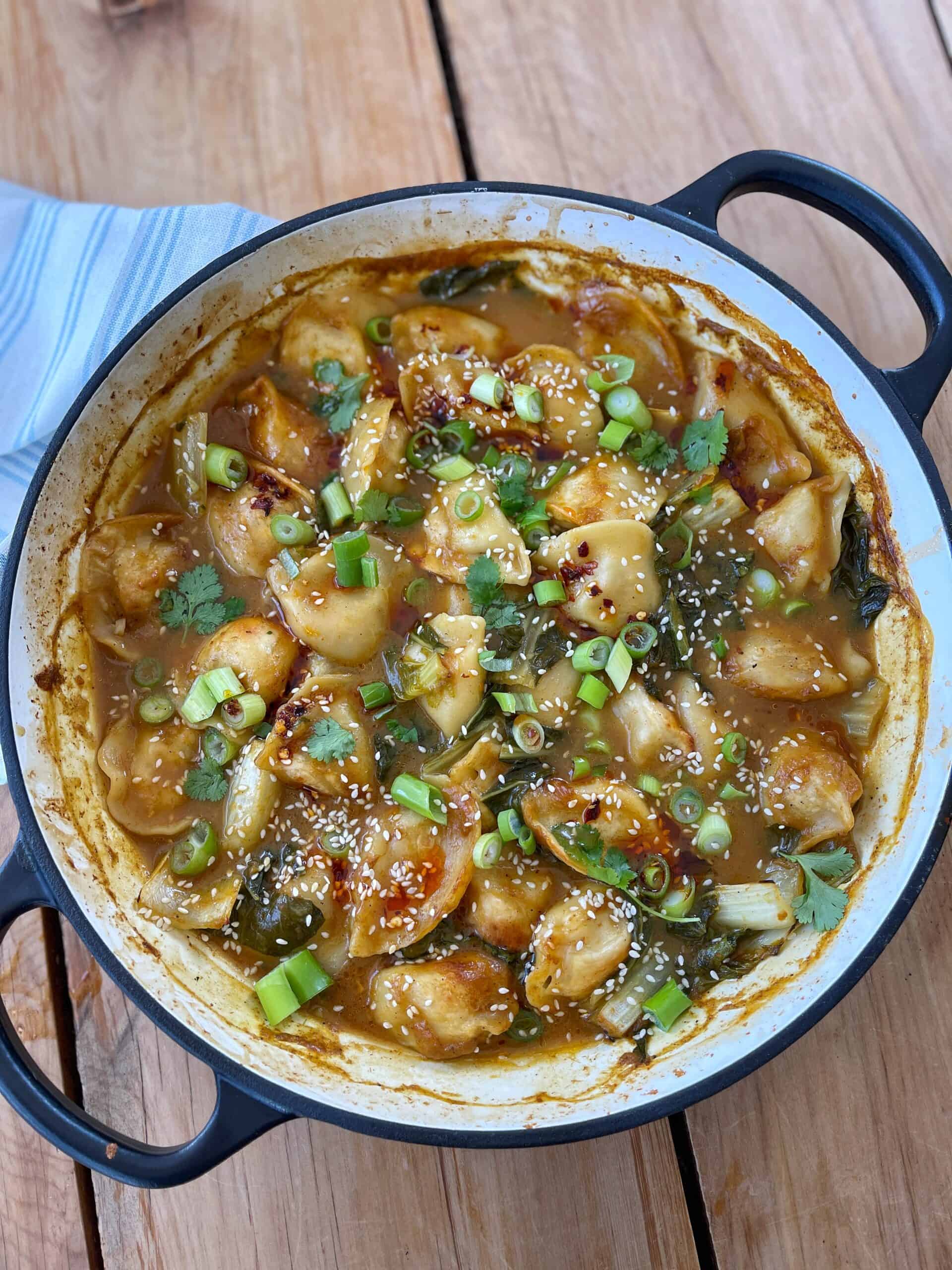 Thai Curry Dumpling Bake A pan filled with dumplings in a brown sauce, topped with chopped green onions, cilantro, and sesame seeds, sits on a wooden surface.