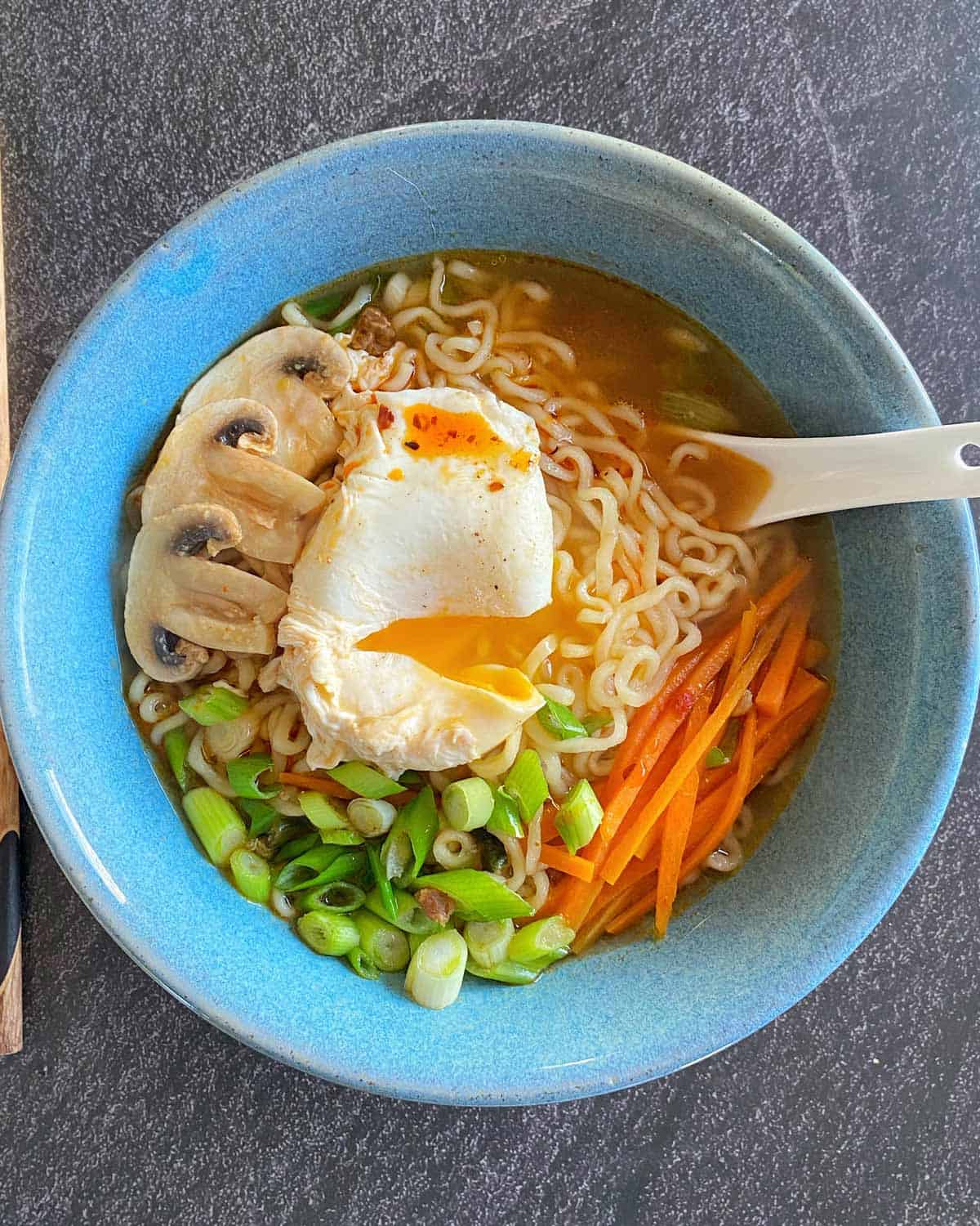 Spicy Ramen and Egg A bowl of ramen topped with a poached egg, sliced mushrooms, green onions, and julienned carrots, with a soup spoon on the side.