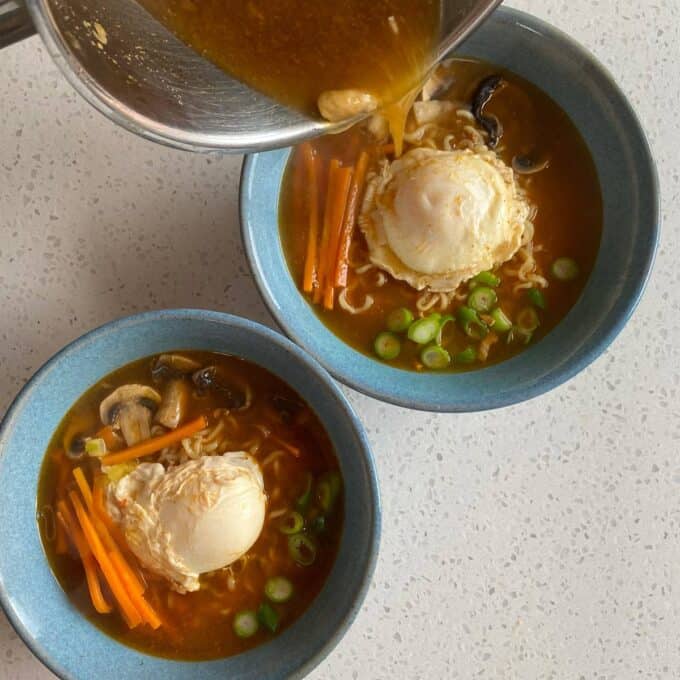 Spicy Ramen and Egg Two blue bowls of ramen with poached eggs, sliced carrots, mushrooms, and green onions; broth is being poured into one of the bowls.