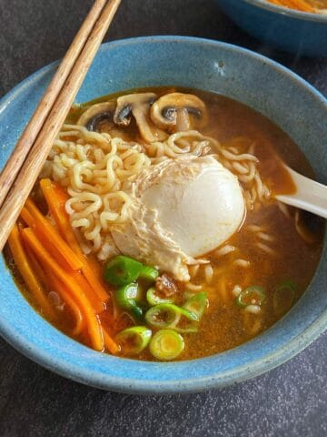 A bowl of ramen with noodles, a poached egg, sliced mushrooms, carrot sticks, and green onions, served with chopsticks and a spoon.