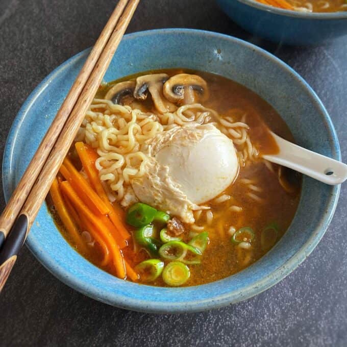 Spicy Ramen and Egg A bowl of ramen with noodles, a poached egg, sliced mushrooms, carrot sticks, and green onions, served with chopsticks and a spoon.
