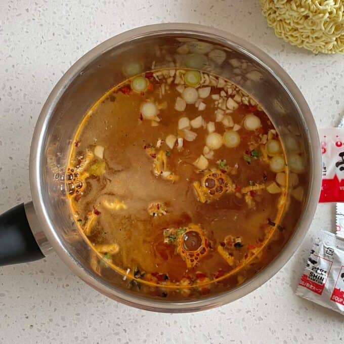Spicy Ramen and Egg A pot of ramen broth with chopped green onions and seasoning visible, sitting on a white countertop next to instant noodle packets and uncooked noodles.