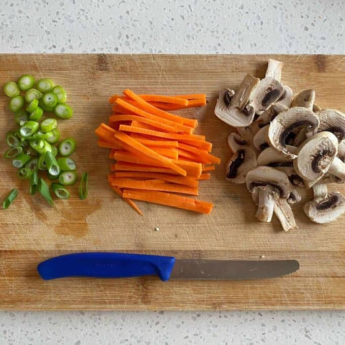 Spicy Ramen and Egg A cutting board with sliced green onions, julienned carrots, and sliced mushrooms next to a knife with a blue handle.