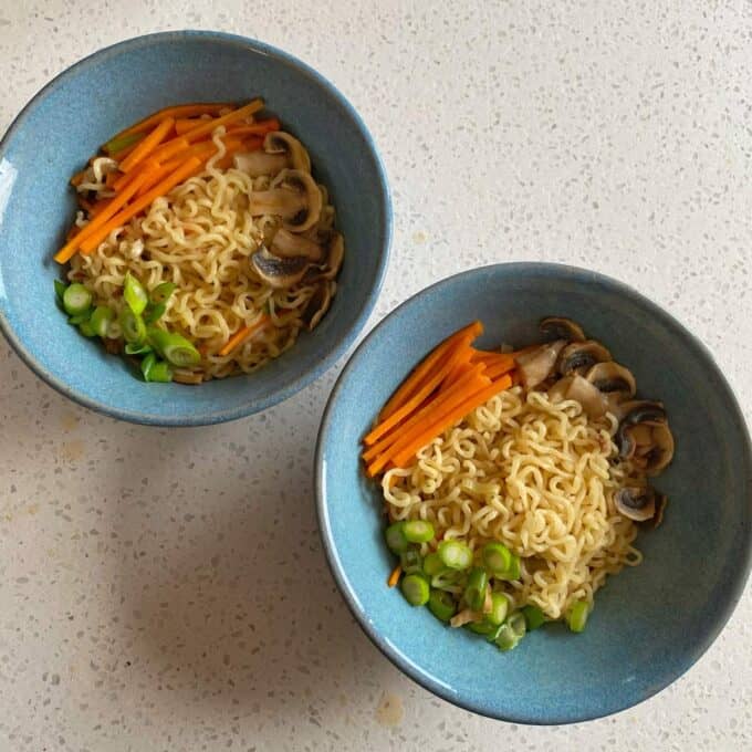 Spicy Ramen and Egg Two blue bowls each containing cooked noodles, sliced carrots, mushrooms, and chopped green onions on a light speckled countertop.