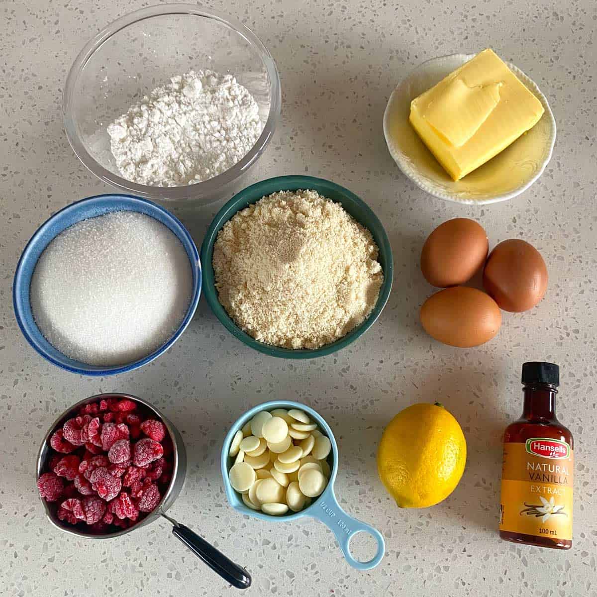 Raspberry Friands A countertop displays flour, butter, eggs, granulated sugar, ground almonds, white chocolate chips, frozen raspberries, a lemon, and vanilla extract in separate bowls and containers.