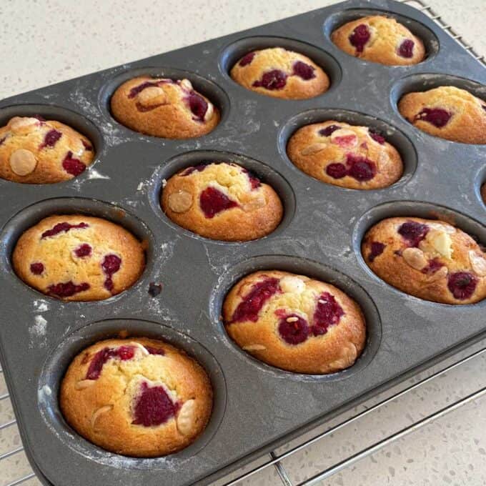 Raspberry Friands A muffin tray on a cooling rack holds twelve baked muffins with visible raspberries and white chocolate chips on top.