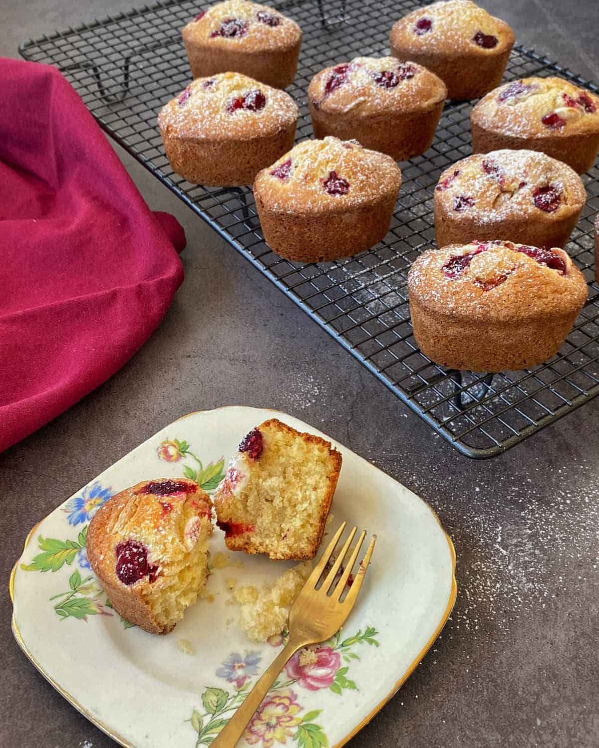Raspberry Friands Eight cherry-topped muffins on a cooling rack, with one muffin cut in half on a floral plate, next to a gold fork and a red cloth on a dark countertop.