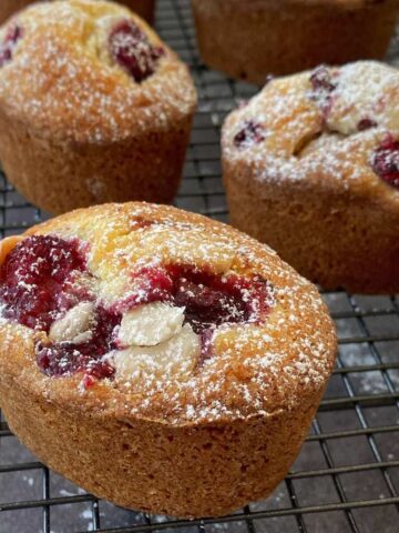 Four golden-brown Raspberry Friands, dusted with powdered sugar, are cooling on a black wire rack.
