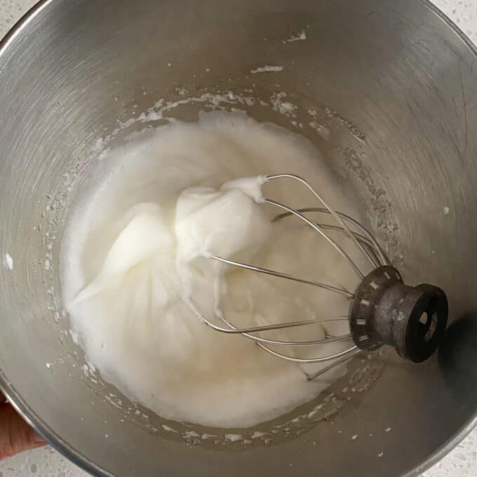 Raspberry Friands A metal mixing bowl contains whipped egg whites with soft peaks, and a wire whisk attachment rests inside the bowl.