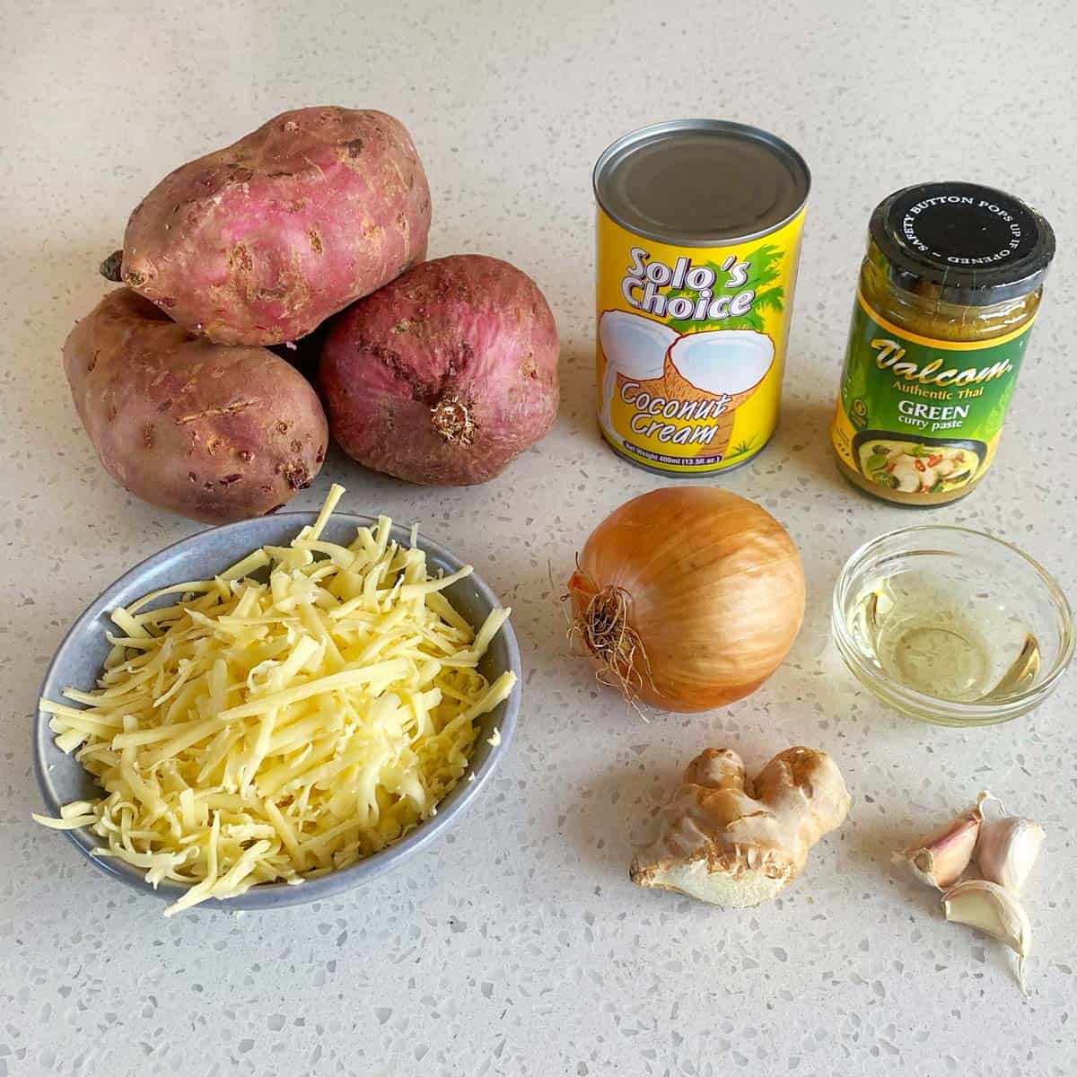 Thai Green Curry Kumara Gratin Sweet potatoes, shredded cheese, a can of coconut cream, green curry paste jar, an onion, oil in a small dish, ginger root, and two garlic cloves arranged on a countertop.