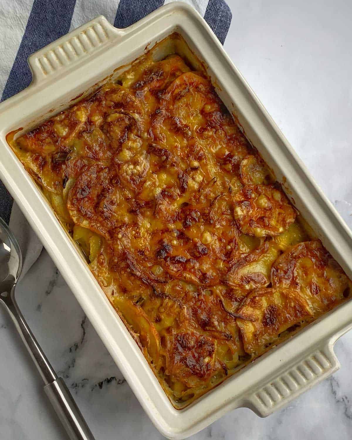Thai Green Curry Kumara Gratin Rectangular baking dish filled with baked cheesy potato gratin, golden brown on top, placed on a marble surface beside a metal spoon.