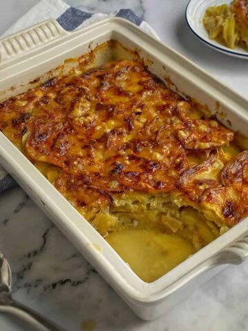 A baked dish of potato gratin with a golden-brown cheese crust, partially served, showing creamy layers and a serving spoon beside the dish.