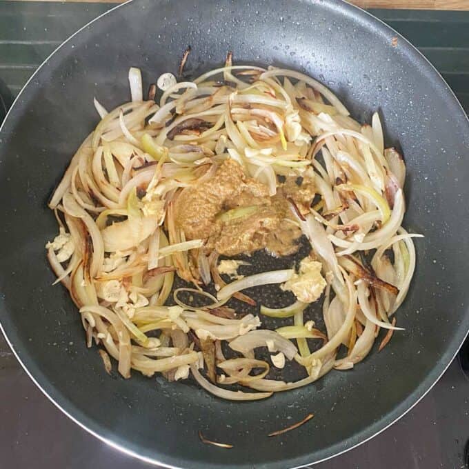 Thai Green Curry Kumara Gratin Sliced onions and garlic are sautéing in a frying pan with a dollop of brown paste in the center.