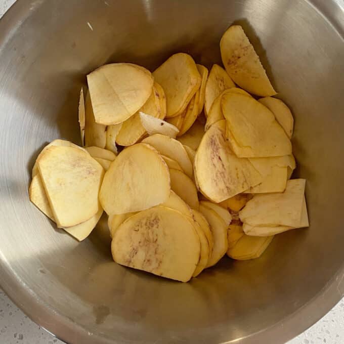 Thai Green Curry Kumara Gratin Thinly sliced raw potatoes are placed in a large stainless steel mixing bowl.