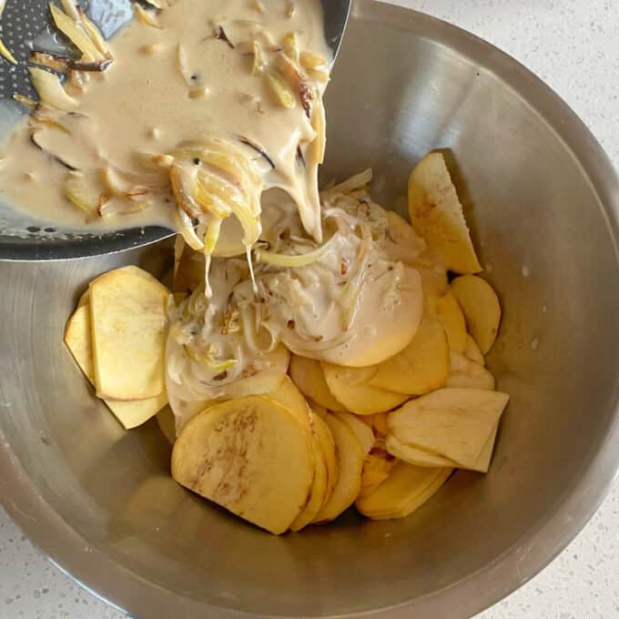 Thai Green Curry Kumara Gratin Sliced potatoes in a metal bowl being covered with a creamy onion mixture poured from a pan.