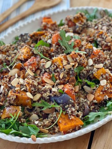 A platter of quinoa salad with roasted sweet potatoes, arugula, almonds, and red onion is served on a wooden table.