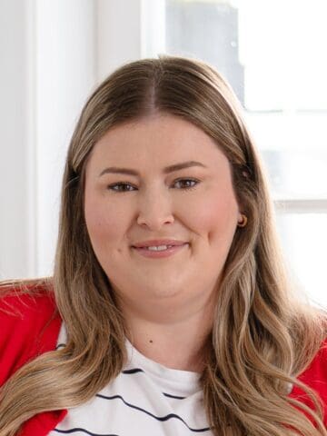 A woman with long, light brown hair wearing a red cardigan and a white striped shirt is smiling in a brightly lit room with a window in the background.