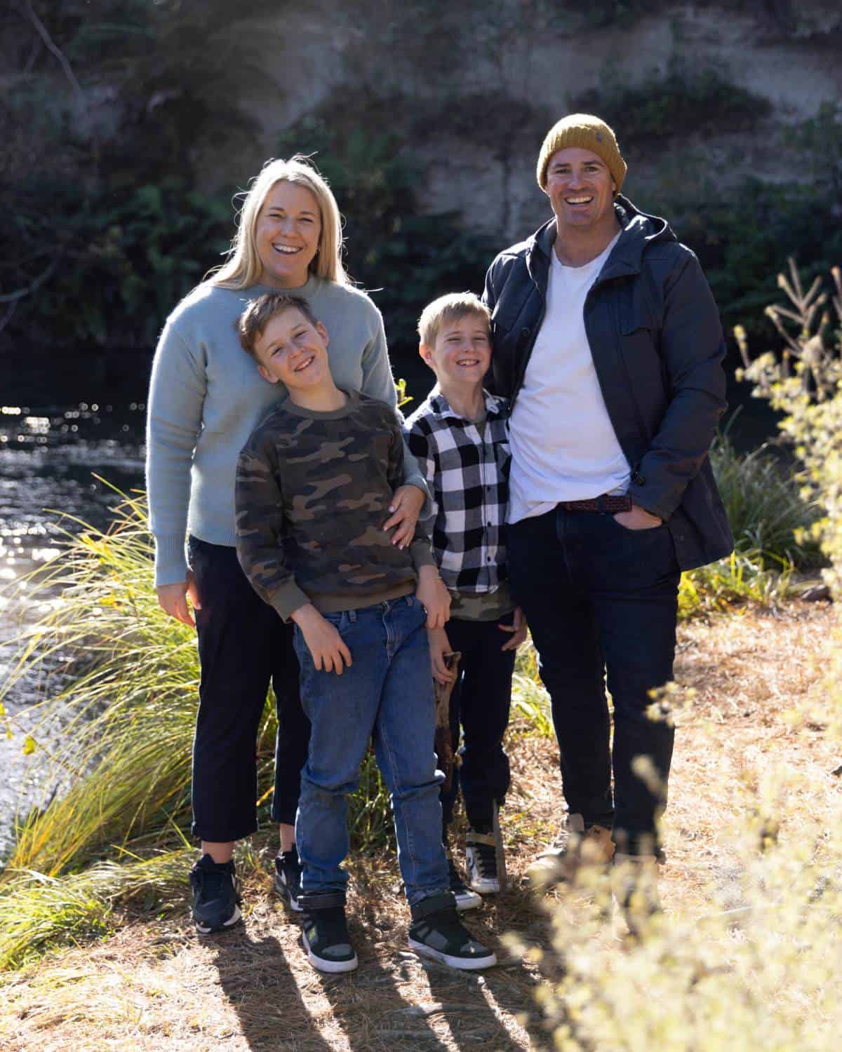 A family of four stands outdoors near a river, smiling at the camera. The adults stand behind two children; all are dressed in casual, warm clothing.