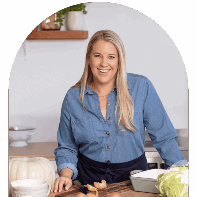A woman in a blue shirt stands in a kitchen, smiling, with onions, a cabbage, and a white pumpkin on the counter in front of her.