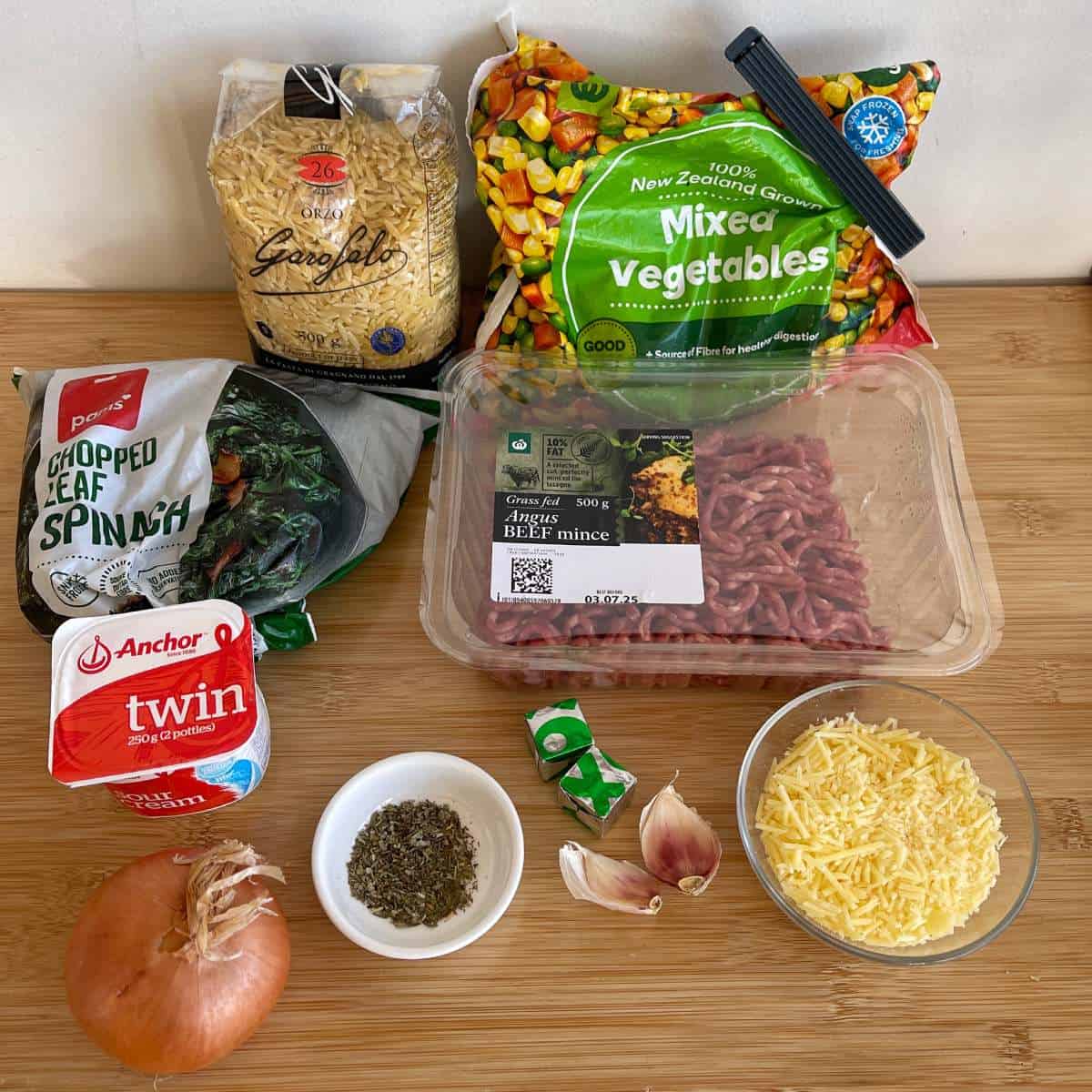 Ingredients for a pasta dish laid on a wooden surface, including pasta, mixed vegetables, spinach, beef mince, cheese, onion, cream, herbs, bouillon cubes, and garlic cloves.