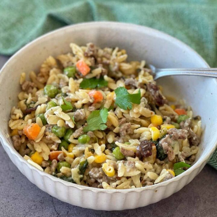 A bowl of pasta mixed with ground meat, peas, corn, carrots, and herbs, with a fork resting inside the bowl.