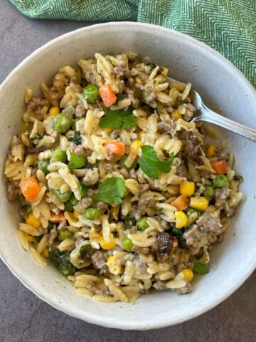 A bowl of creamy orzo pasta mixed with ground meat, peas, corn, carrots, and herbs, with a fork resting inside the bowl.