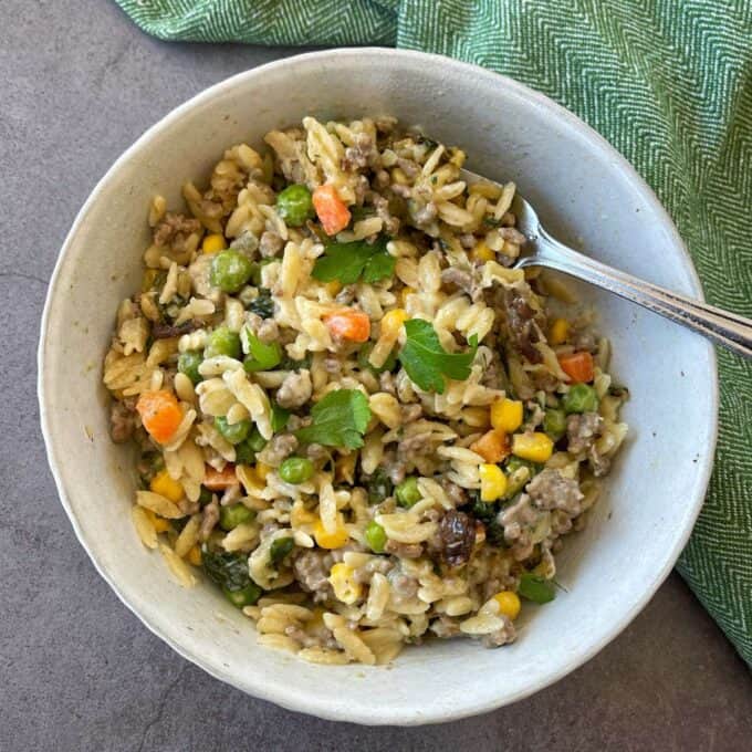 A bowl of creamy orzo pasta mixed with ground meat, peas, corn, carrots, and herbs, with a fork resting inside the bowl.