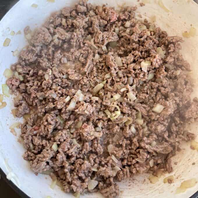 Ground beef and chopped onions are being cooked together in a white skillet.