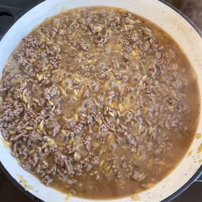 Ground beef, orzo pasta, and onions simmer in a brown broth inside a large white skillet on a stovetop.
