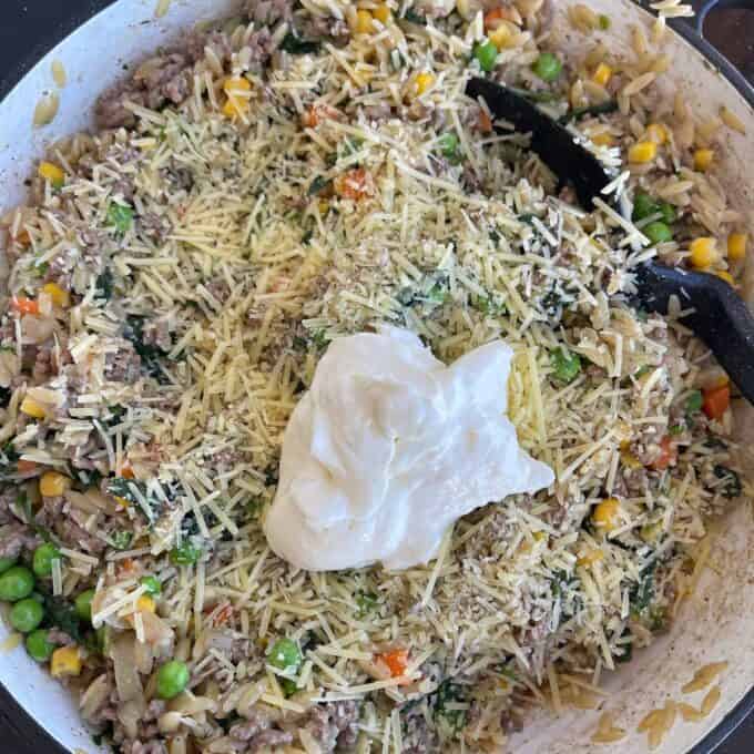 A skillet containing a mixture of ground meat, rice, peas, corn, shredded cheese, and a dollop of sour cream, with a black spatula on the side.